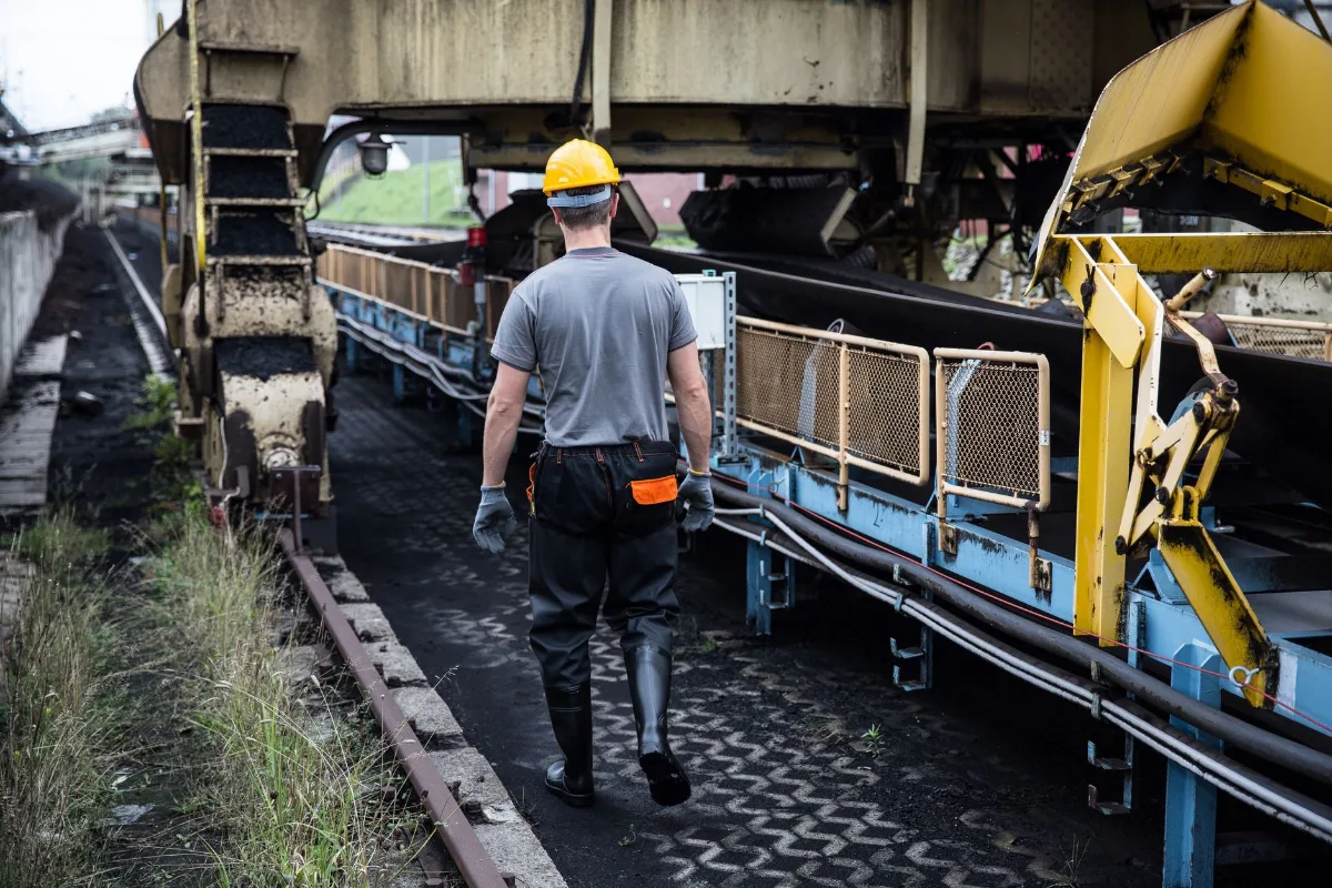 Trabalhador de capacete amarelo caminhando de costas ao lado de uma correia transportadora numa planta industrial
