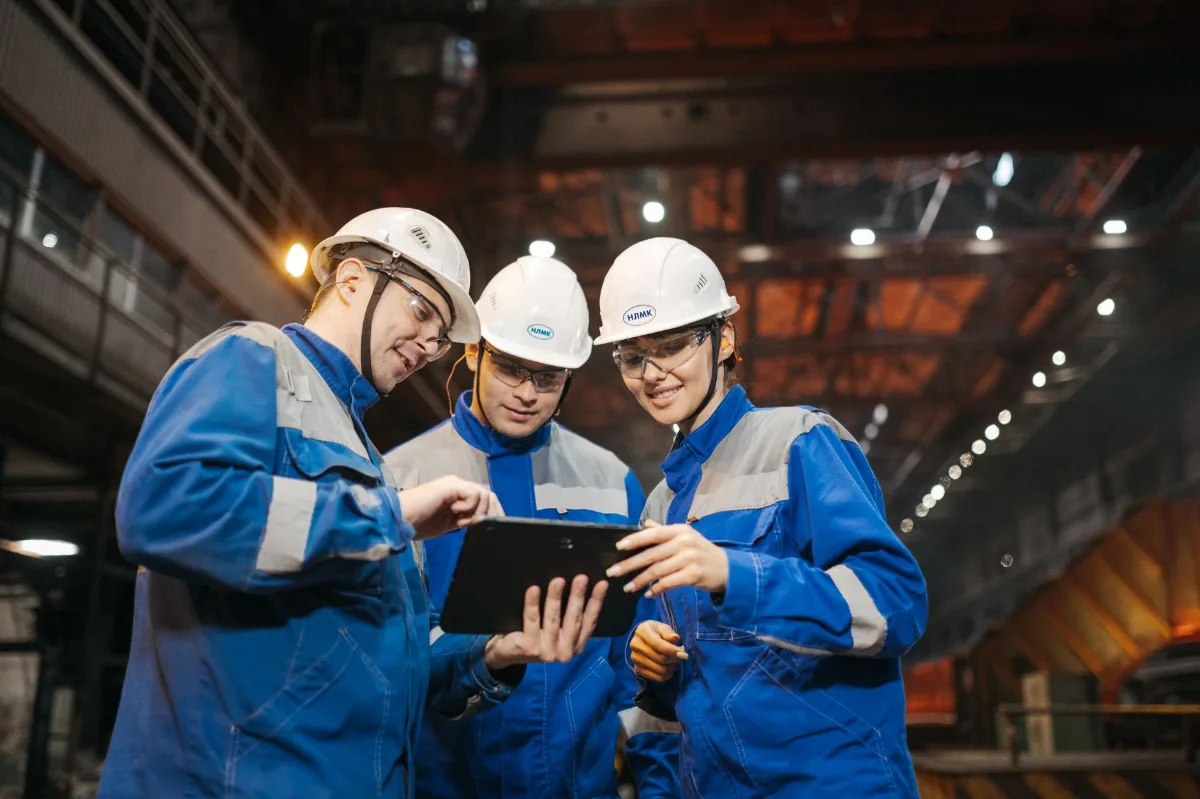 Três engenheiros de capacete branco, óculos de segurança e uniforme azul analisam dados em um tablet dentro de uma usina siderúrgica