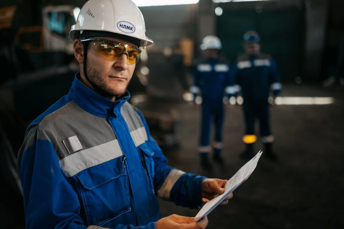 Profissional de capacete branco e óculos de proteção segurando documentos técnicos num galpão industrial, com colegas de uniforme ao fundo