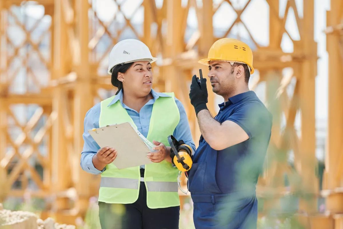 Dois engenheiros com capacete e colete de segurança conversando em canteiro de obra, com andaimes ao fundo