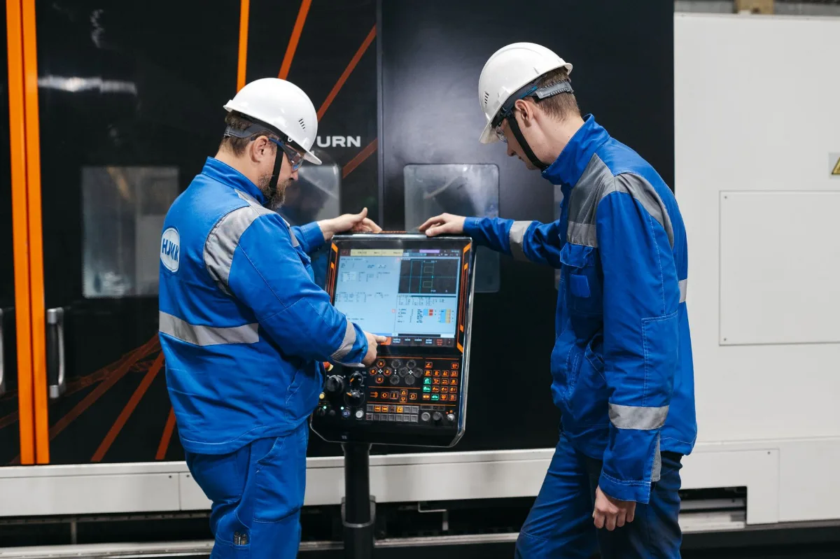 Dois engenheiros de capacete e óculos de segurança operando o painel de controle de um torno CNC em uma fábrica