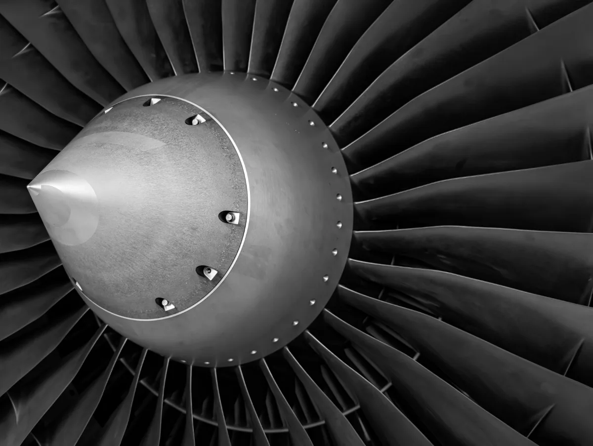 Fotografia em preto e branco, em close, do fan de um motor turbofan de avião, com o cone central aparafusado e as pás metálicas curvas em volta