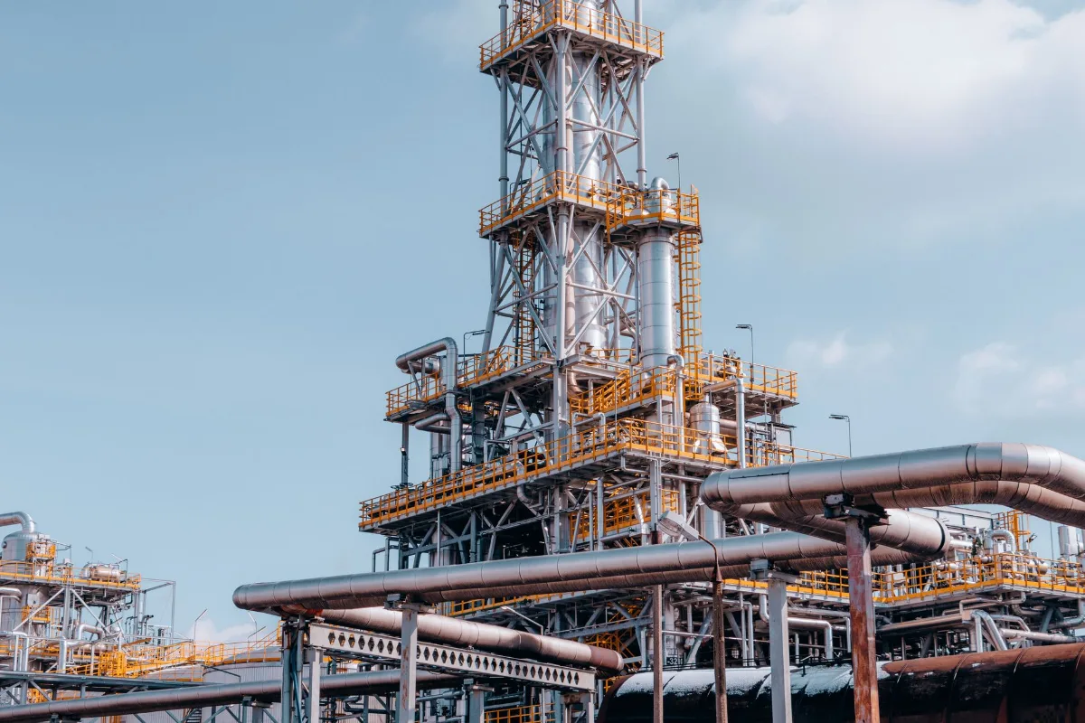 Vista externa de uma refinaria de petróleo com torres de destilação, tanques e uma rede densa de tubulações metálicas
