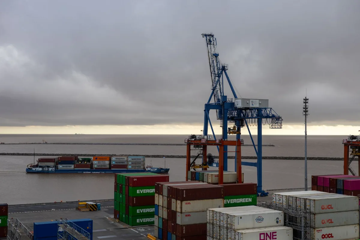 Portêiner azul sobre pórtico laranja em terminal de contêineres, com pilhas de contêineres no pátio e navio porta-contêineres passando no canal sob céu nublado
