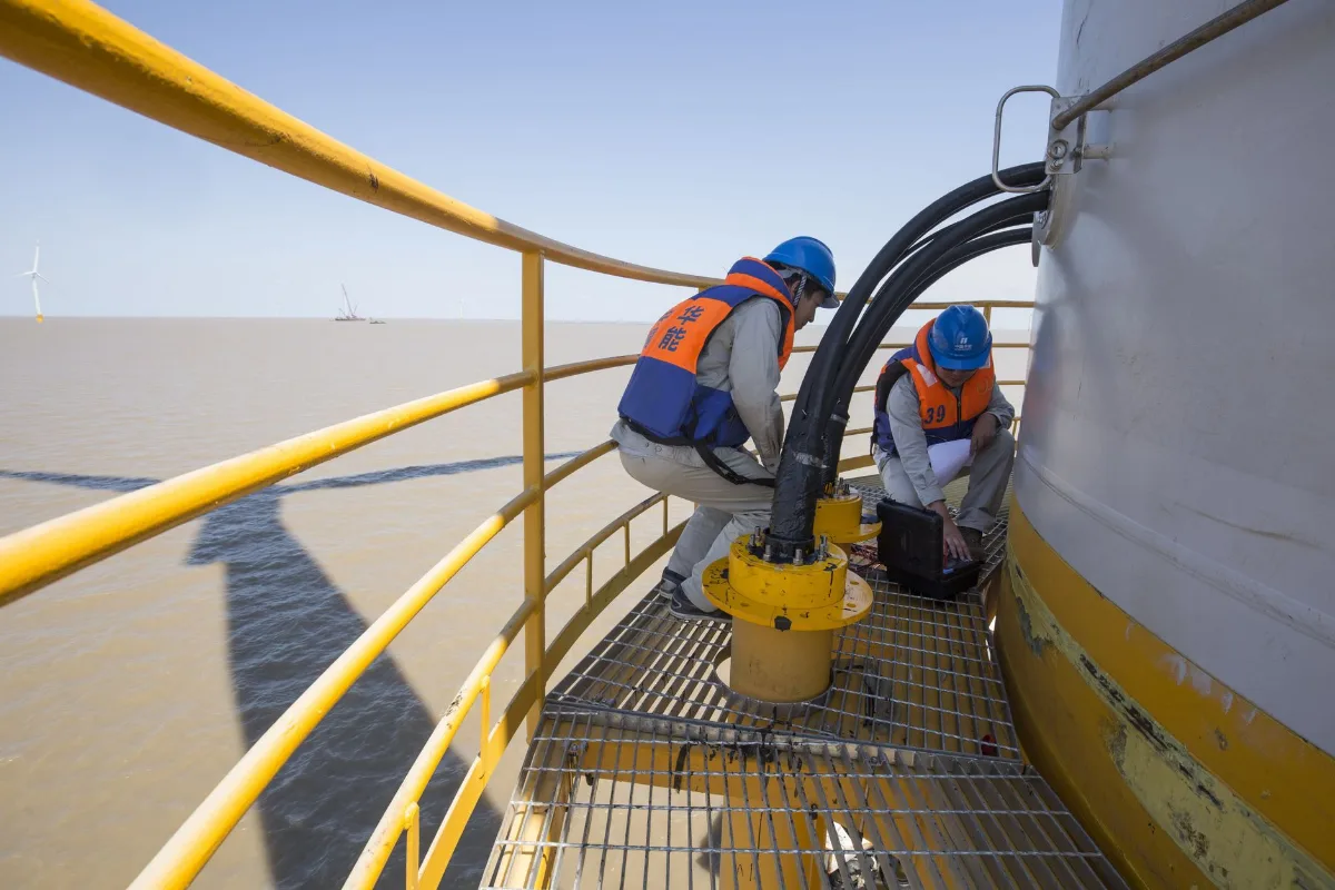 Técnicos com capacete, cinto e equipamento de segurança fazendo manutenção no alto de uma turbina eólica offshore, com o mar ao fundo