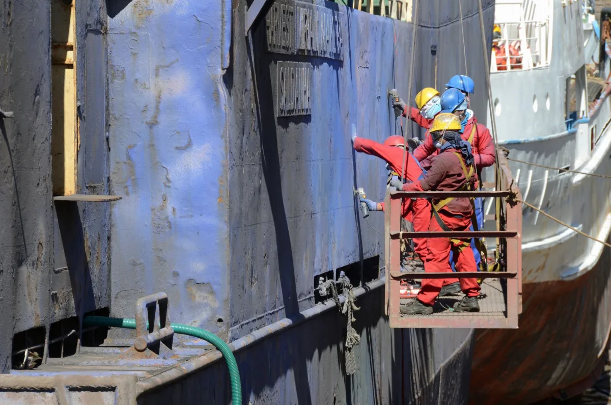 Operários com capacete, macacão vermelho e cinto de segurança em plataforma suspensa preparando o casco de um navio em reparo no estaleiro