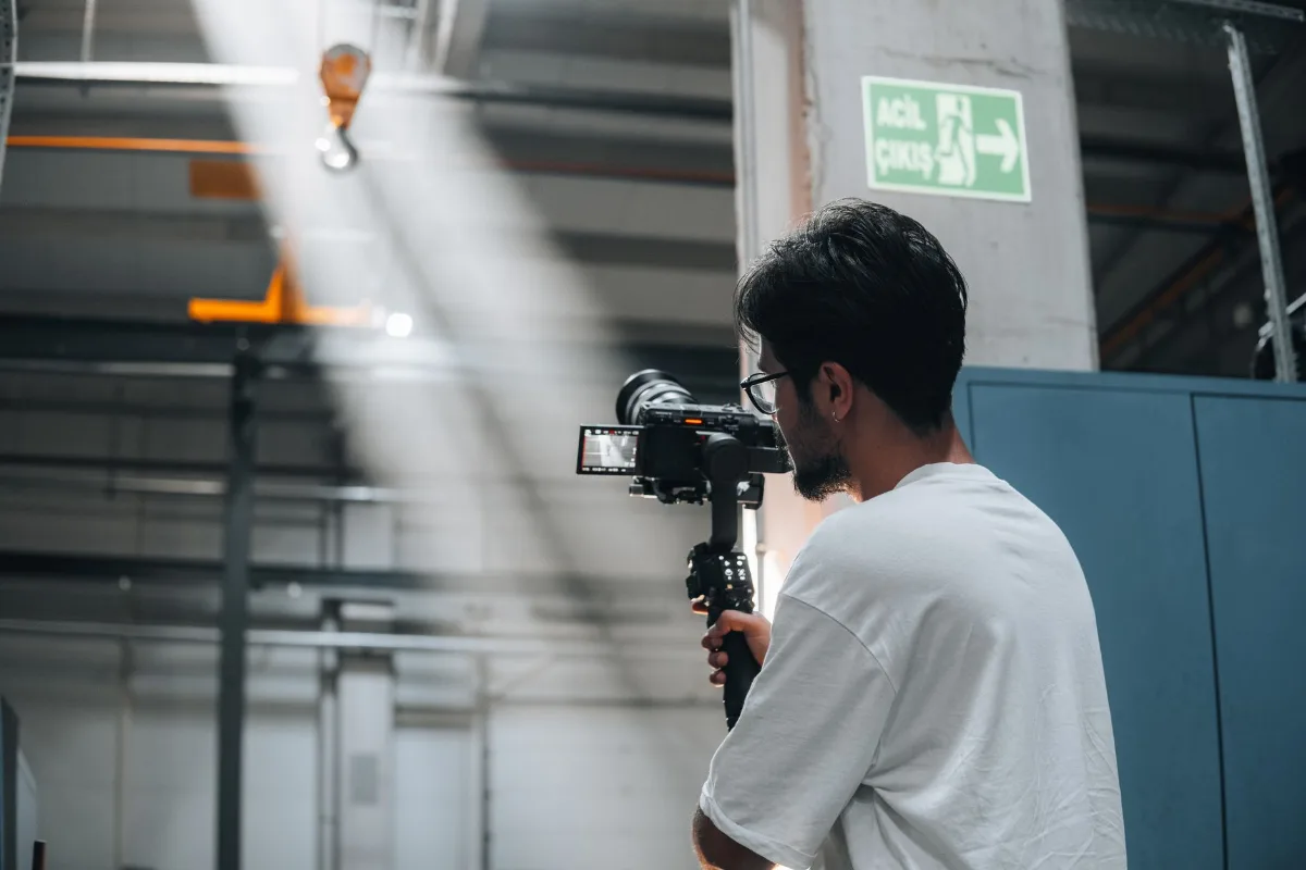 Cinegrafista com câmera em estabilizador filmando o interior de um galpão industrial com ponte rolante e feixe de luz