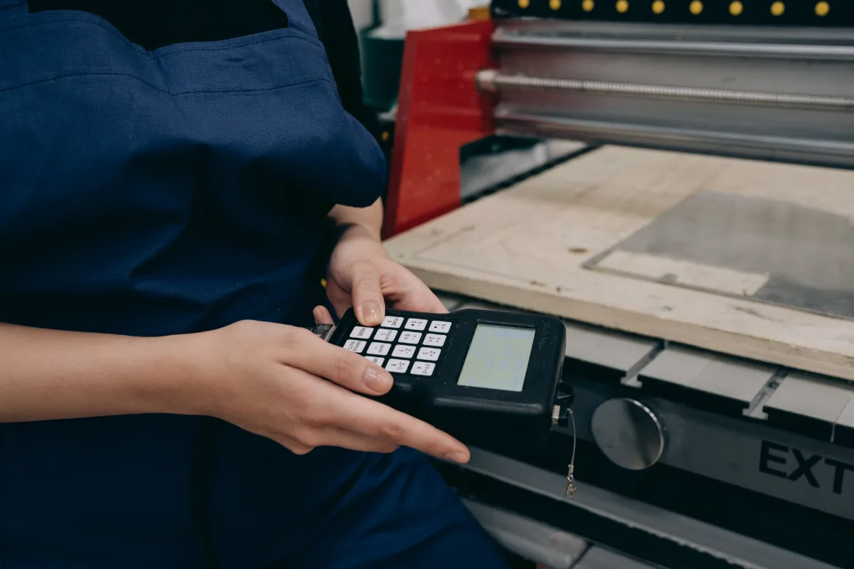 Operadora de uniforme azul programando uma máquina CNC com controlador manual ao lado da mesa de corte