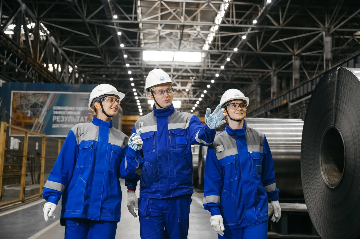 Três profissionais de uniforme azul e capacete caminhando entre bobinas de aço numa usina, um deles apontando para a linha