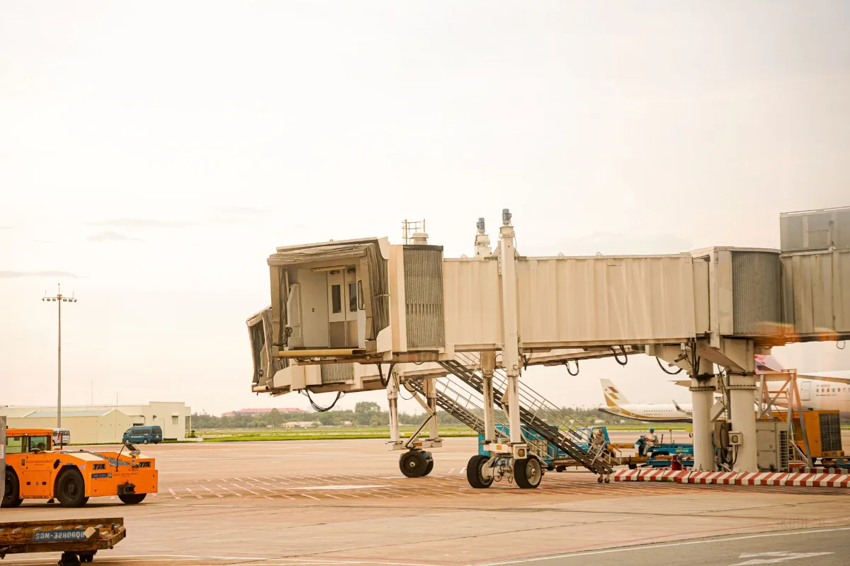 Ponte de embarque recolhida no pátio de um aeroporto, com rodado de apoio, escada de serviço, trator rebocador laranja e equipamentos de solo ao lado