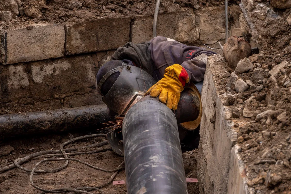 Soldador com máscara de solda e luvas de raspa amarelas unindo um tubo de aço revestido dentro de uma vala aberta, com paredes de blocos de concreto, terra escavada e cabos de solda no fundo