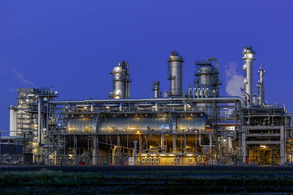 Planta de processo química iluminada ao entardecer, com colunas de destilação, vasos horizontais de grande porte, racks de tubulação metálica e escadas de acesso sob céu azul