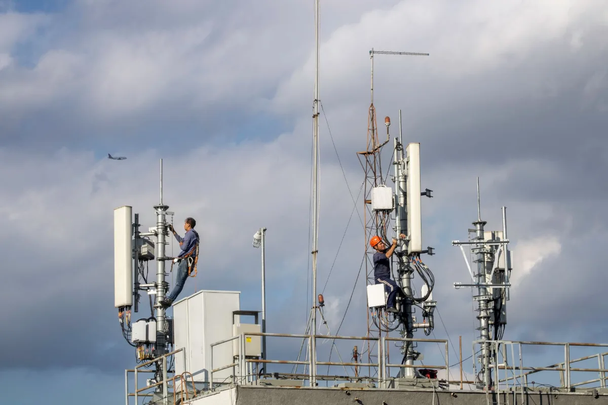 Dois técnicos com cinto de segurança fazem manutenção em antenas de telefonia celular instaladas no topo de um prédio, sob céu nublado
