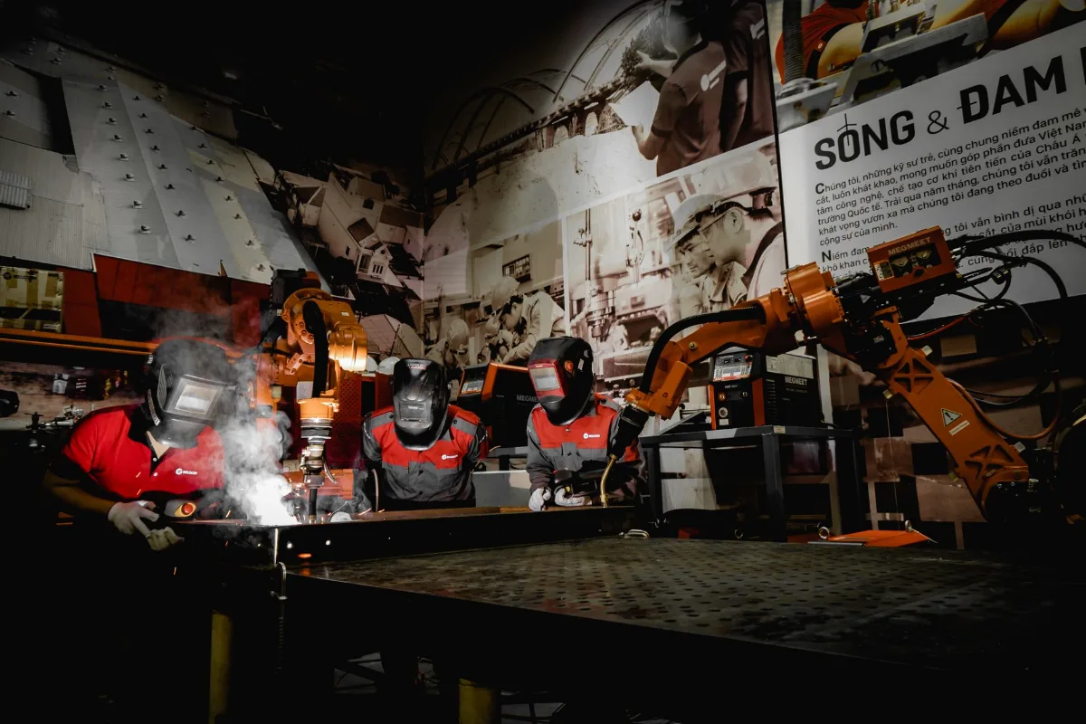Três soldadores com máscara de solda, luvas e uniforme de proteção trabalham em uma bancada metálica ao lado de braços robóticos de soldagem laranja em uma oficina industrial