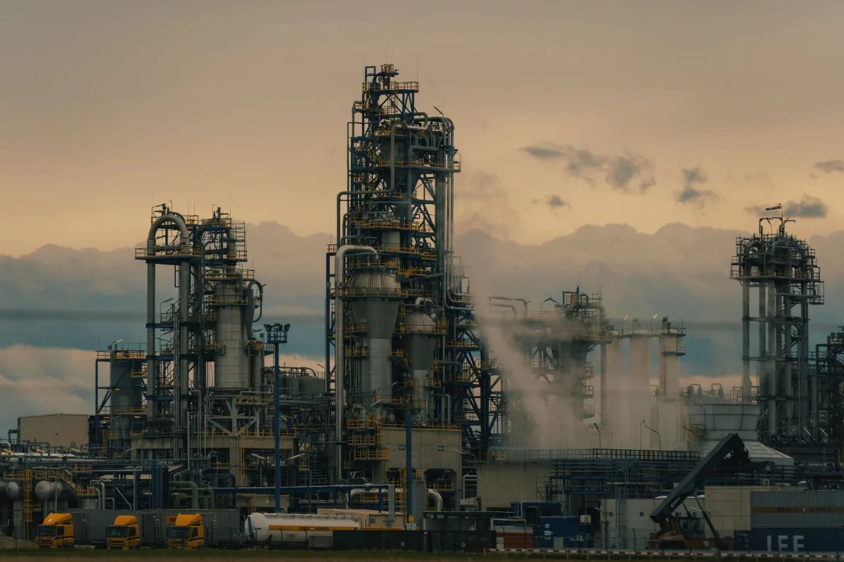 Torres de destilação e tubulações de uma refinaria ao entardecer, com vapor saindo das unidades e caminhões estacionados em primeiro plano