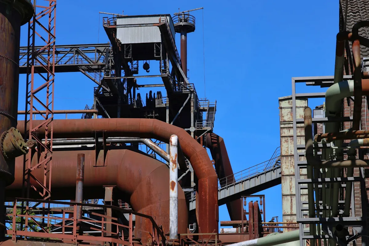 Estrutura de usina com tubulações de aço enferrujadas, passarelas metálicas, escadas e torre de equipamento sob céu azul