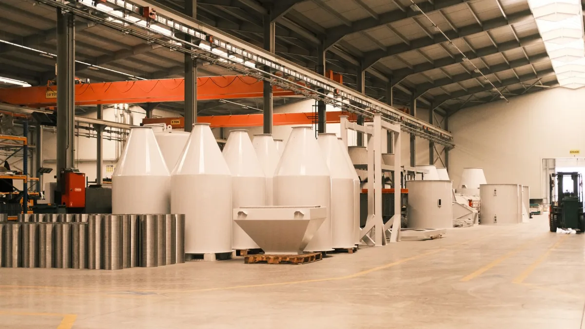 Galpão de fabricação metálica com pontes rolantes laranja, silos e tremonhas de aço pintados de branco sobre paletes, bobinas de chapa e empilhadeira ao fundo