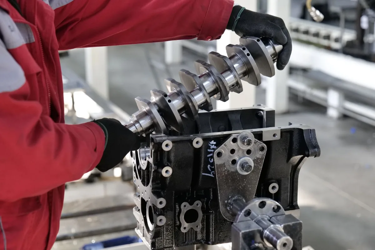 Operador de uniforme vermelho e luvas pretas encaixa um virabrequim usinado em um bloco de motor preso a um dispositivo de montagem, em linha de produção industrial