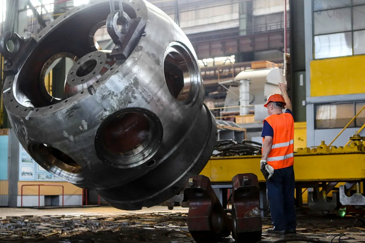 Peça metálica pesada de grande diâmetro içada por cabos em um galpão industrial, observada por um operário de capacete laranja e colete refletivo